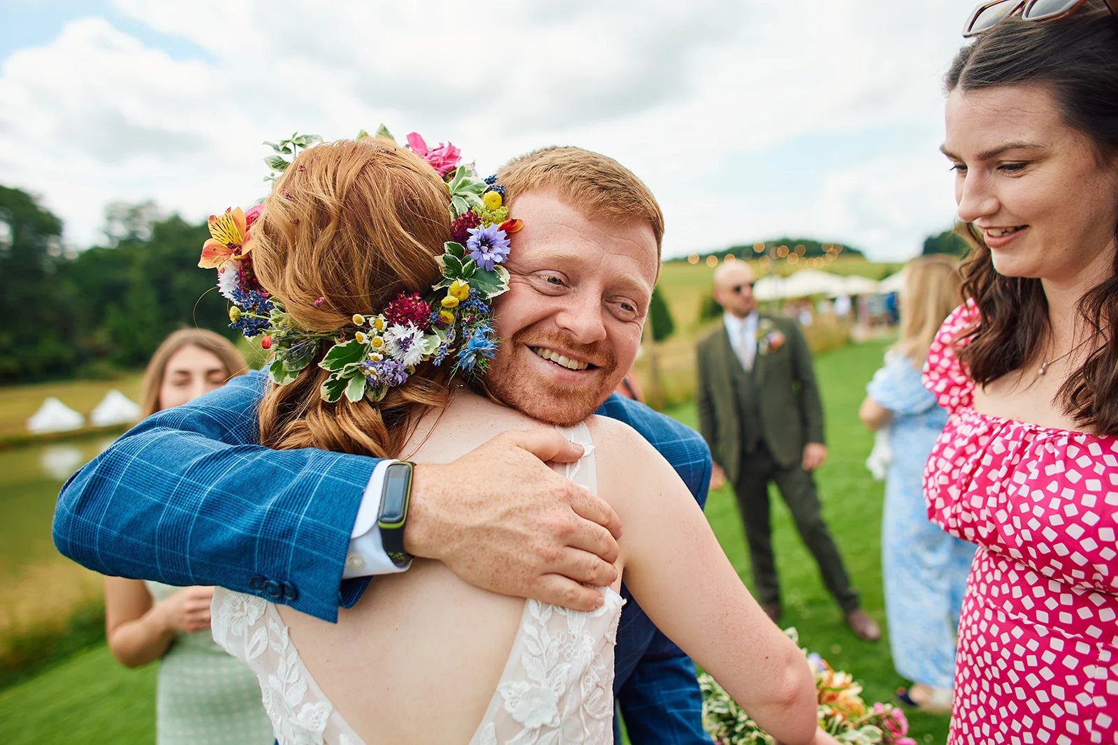 A joyful groom wearing a checked suit embraces his bride, who is adorned with a vibrant fresh flower crown during an outdoor summer wedding reception.