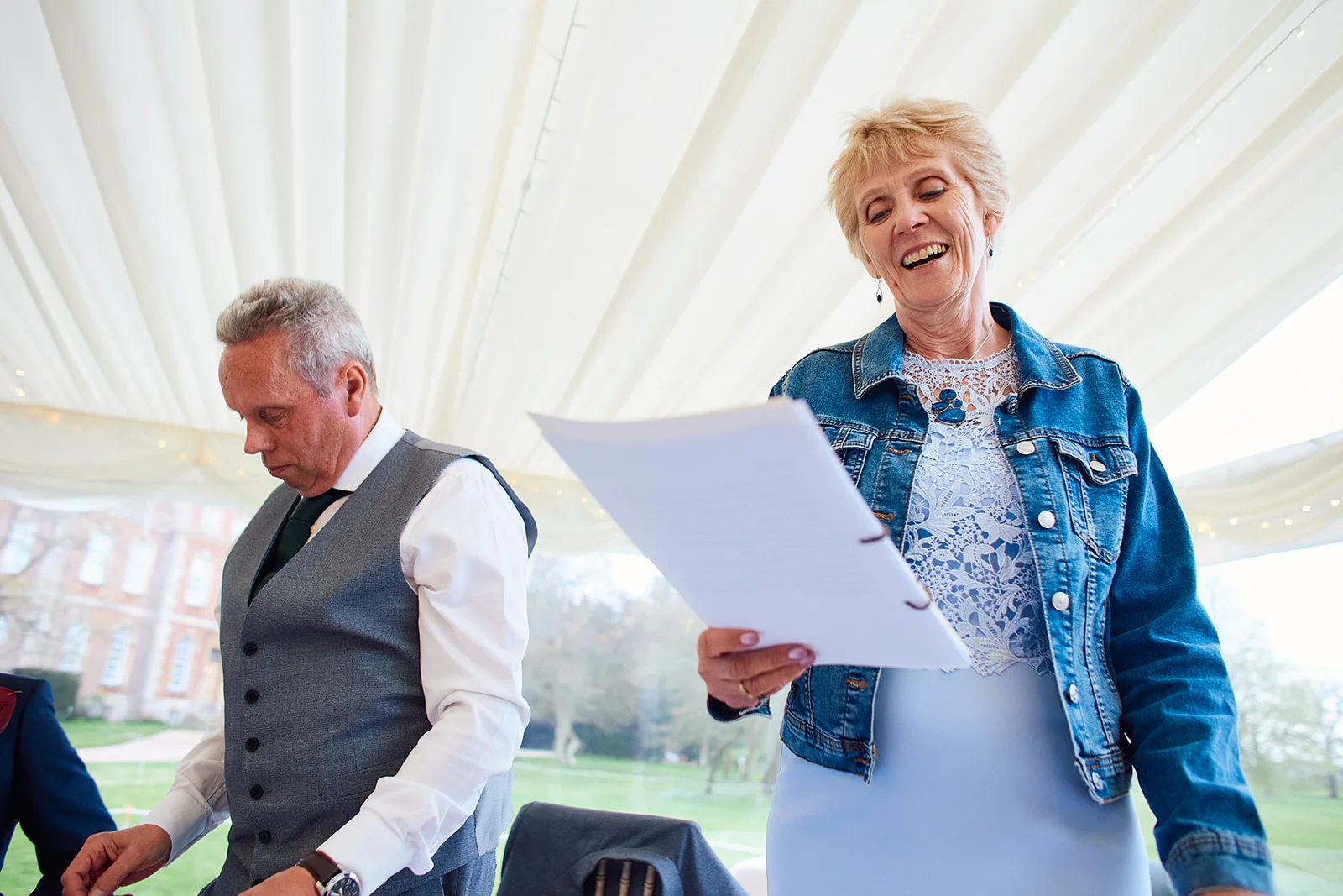 A smiling mother of the bride joyfully delivers her speech holding papers inside a draped marquee while a gentleman looks down beside her.
