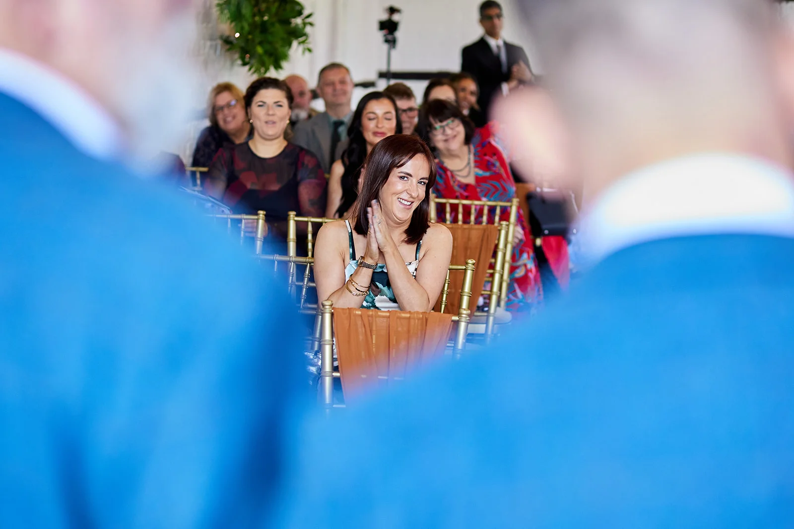 A smiling wedding guest claps her hands happily while seated amongst a gathering of friends and family watching the ceremony.