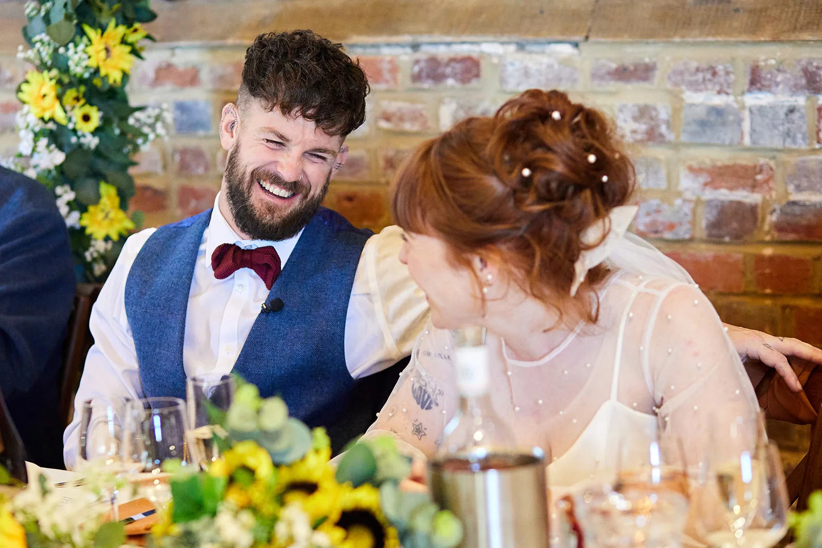 A newlywed couple shares a joyful belly laugh together at the top table during their rustic barn wedding reception, surrounded by sunflowers and exposed brick.