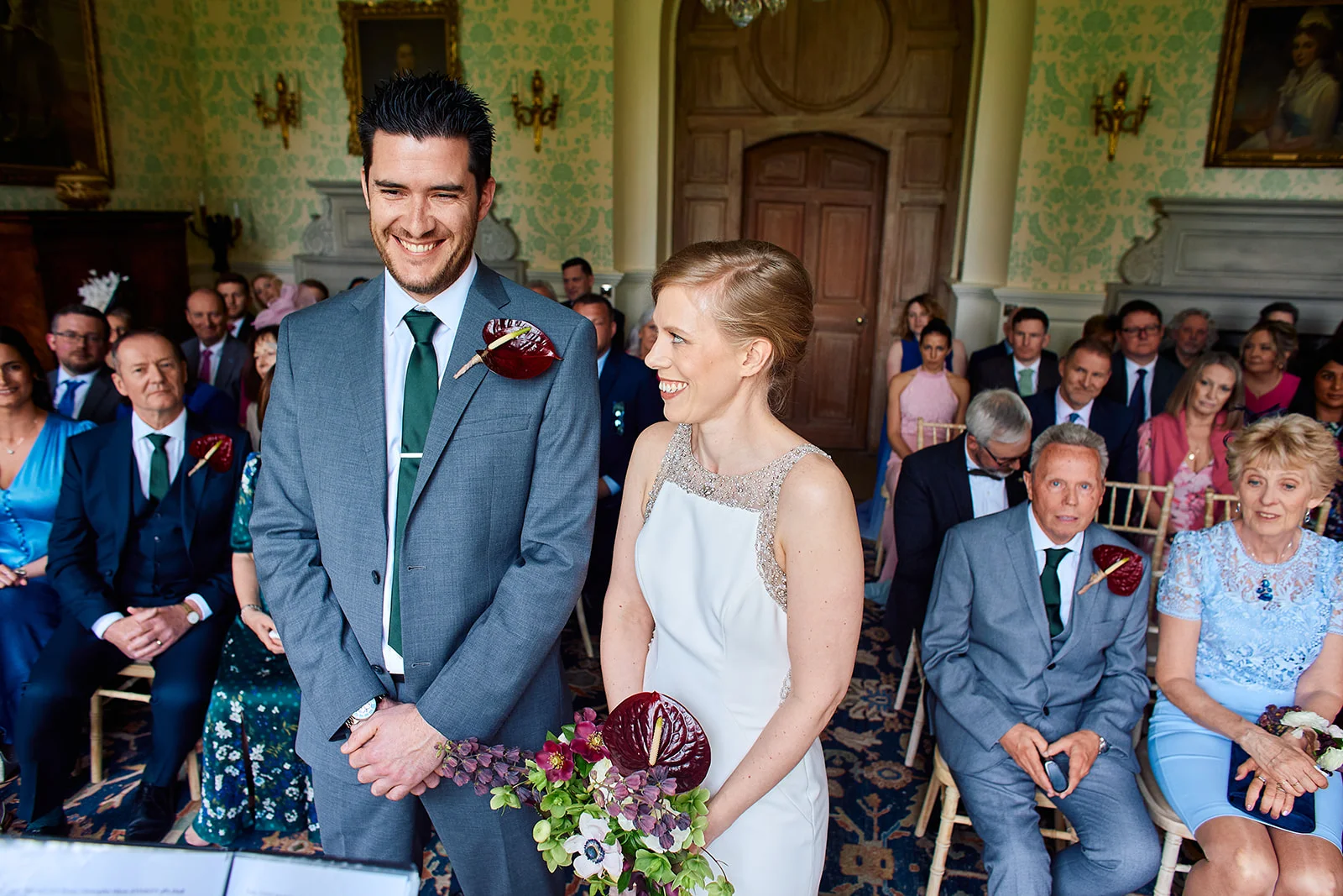 A smiling bride and groom stand together at the front of a panelled ceremony room surrounded by seated wedding guests.