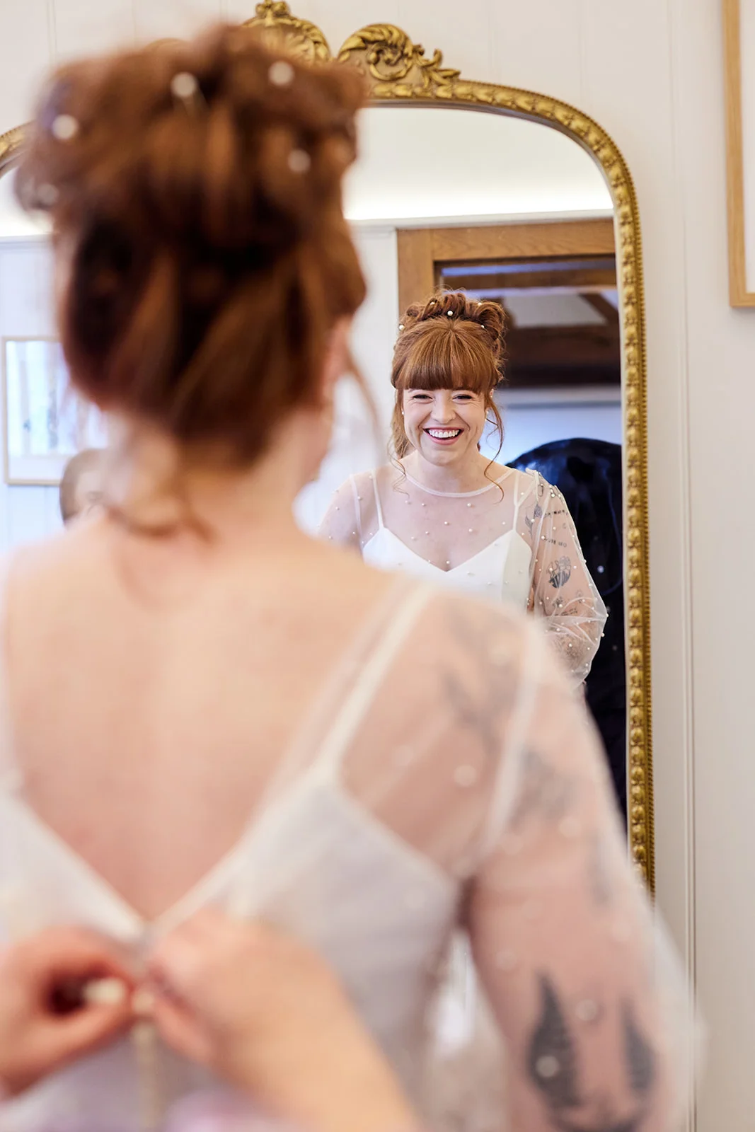 A smiling red-haired bride looks into an ornate gold mirror while her wedding dress is being fastened from behind during morning preparations.