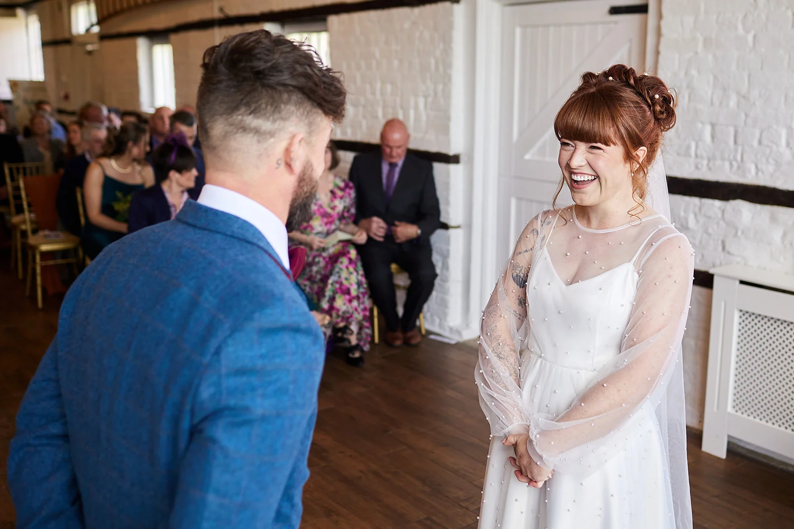 A joyful red-haired bride wearing a pearl-embellished gown laughs happily as she stands at the altar opposite her groom during their indoor barn ceremony.