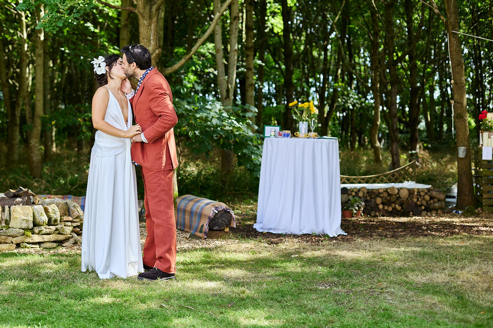 A newlywed couple shares a tender kiss in a sun-dappled woodland setting during an outdoor wedding ceremony.
