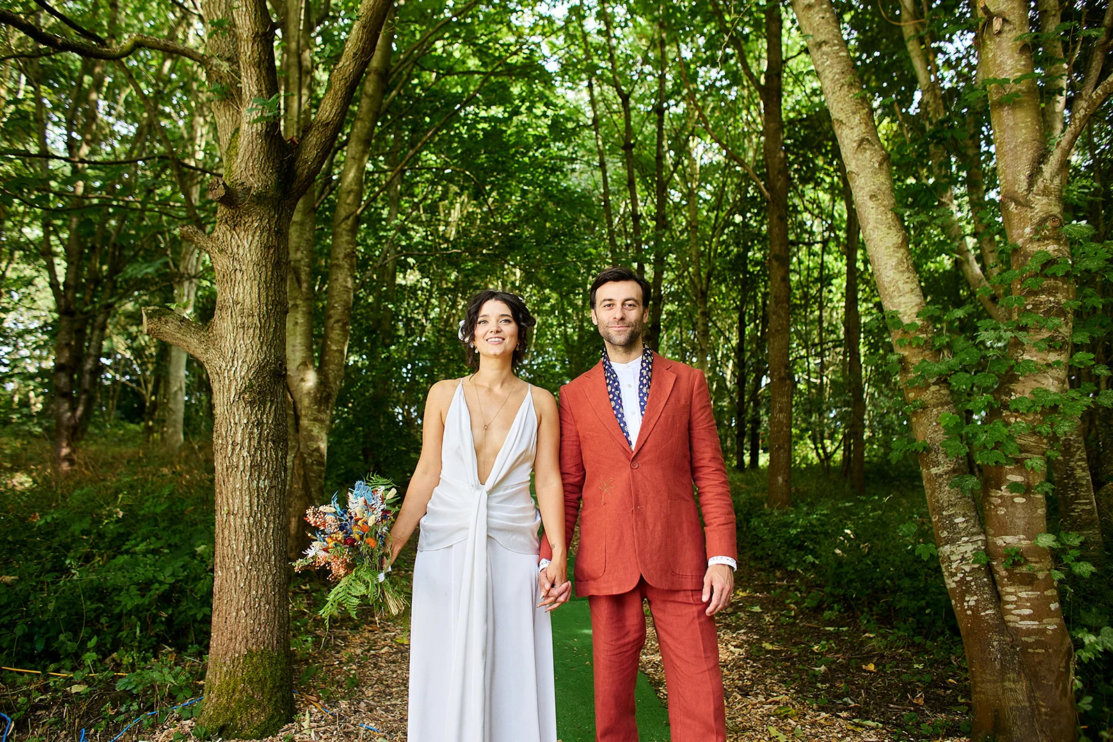 A newlywed couple hold hands while walking along a lush woodland path, with the bride wearing a flowing white v-neck gown and holding a vibrant bouquet, and the groom dressed in a stylish rust-coloured suit.