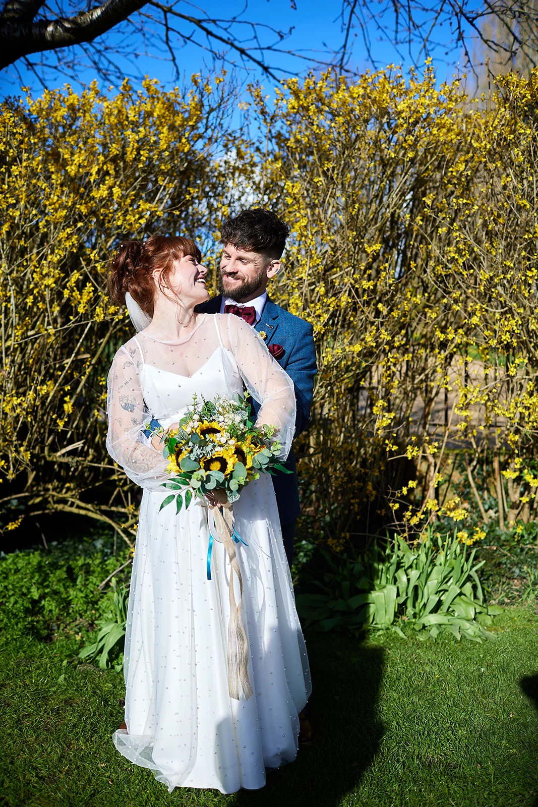 A smiling bride and groom embrace in a sunlit garden, surrounded by vibrant yellow blossoms and lush greenery.