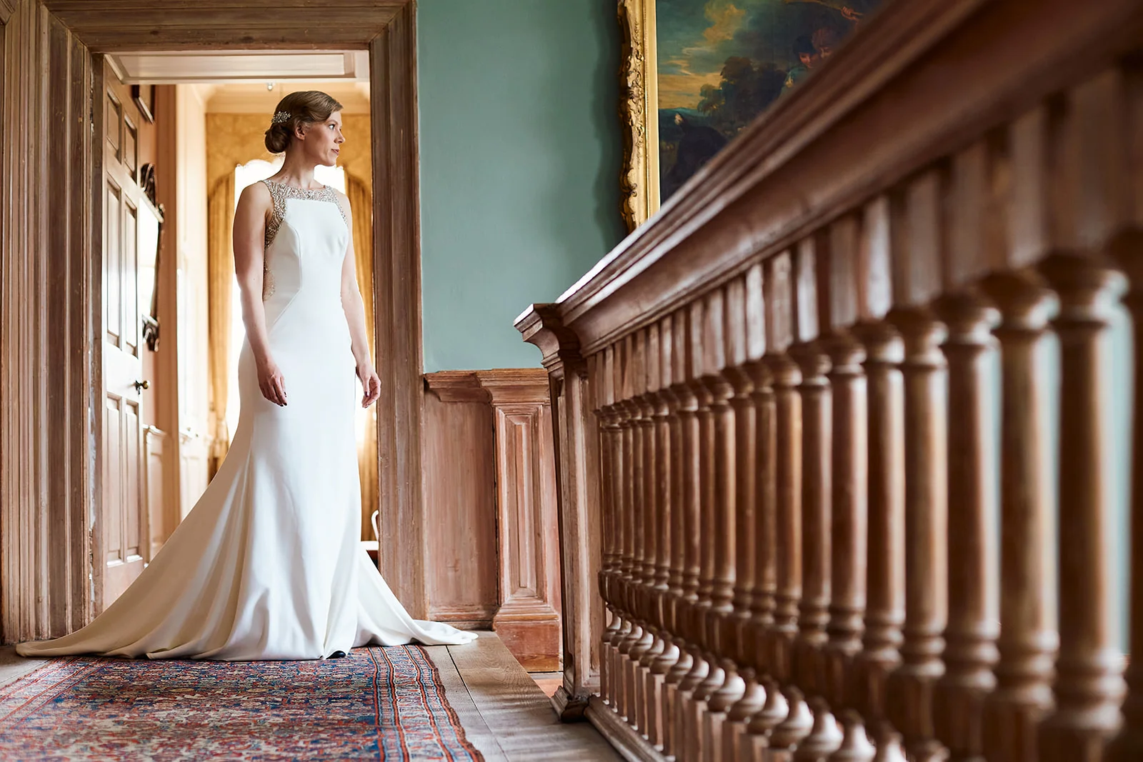 A stunning solo bride poses by a grand wooden staircase in a historic manor house while wearing an elegant high-necked crepe wedding gown.