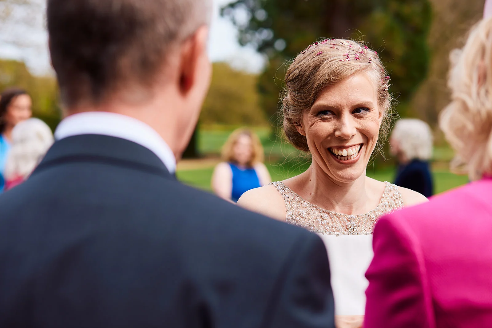A joyful bride laughs openly while interacting with guests outdoors during a wedding reception, captured from an over-the-shoulder perspective.