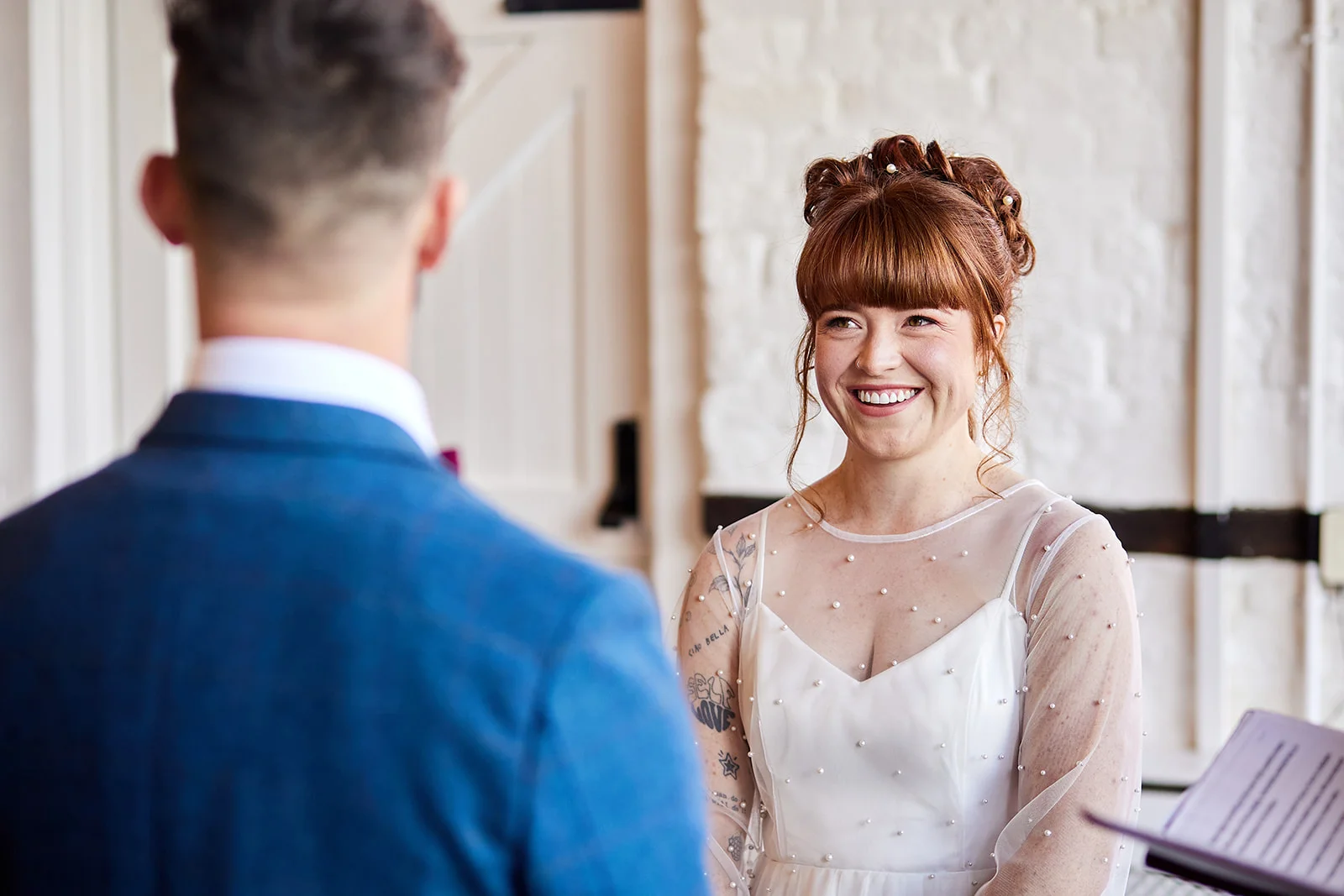 A smiling bride with red hair and a pearl-embellished gown looks joyfully at the groom during their wedding ceremony.