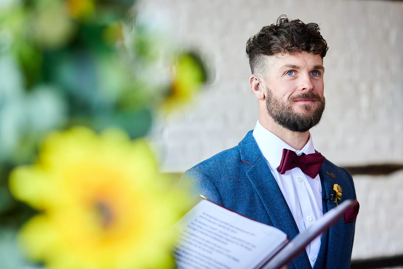 A groom in a blue tweed suit and a burgundy bow tie looks ahead emotionally during the wedding ceremony, framed by blurred yellow sunflowers in the foreground.