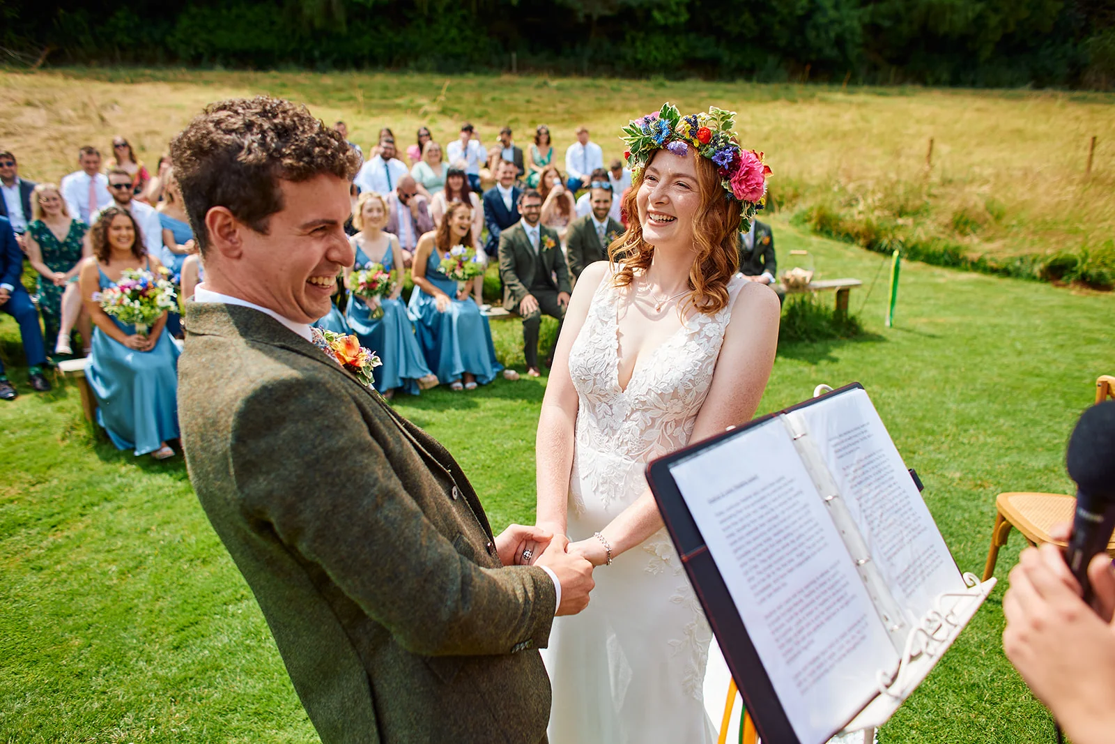 A smiling bride and groom laugh together holding hands during their outdoor wedding ceremony on a bright summer day, watched by seated guests.