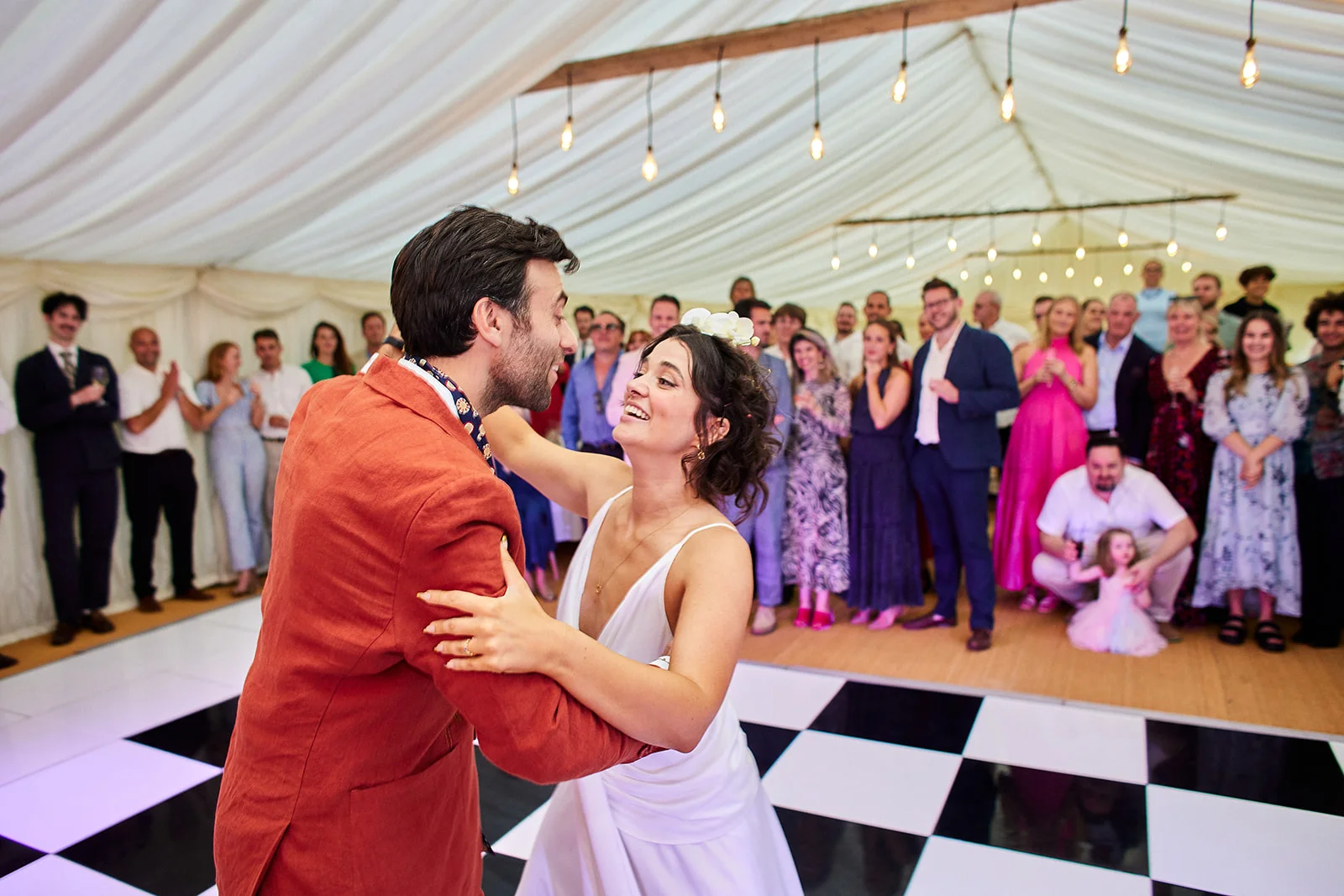 A bride and groom share a joyful first dance on a black and white checkerboard dance floor inside a brightly lit marquee, surrounded by cheering wedding guests.
