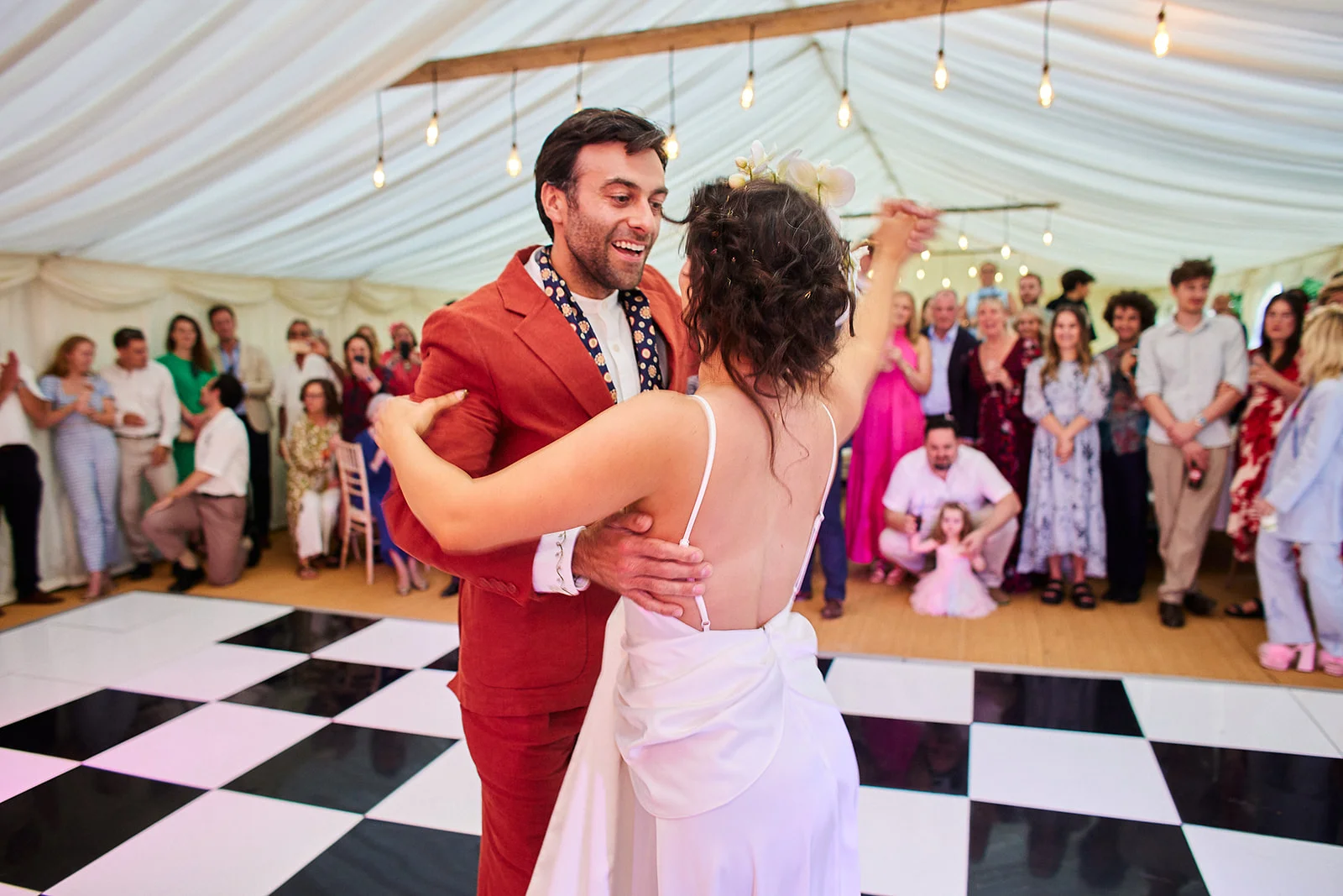 A newlywed couple shares their first dance on a black and white checkered dance floor inside a marquee filled with watching guests.