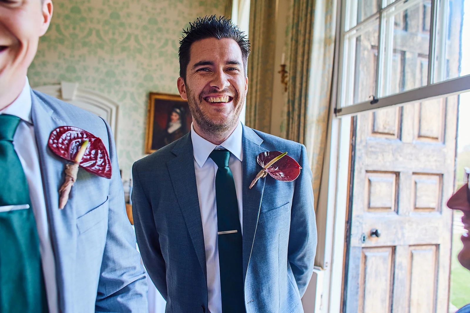 A smiling groom wearing a grey suit and dark green tie with an anthurium buttonhole stands indoors near a wooden door.