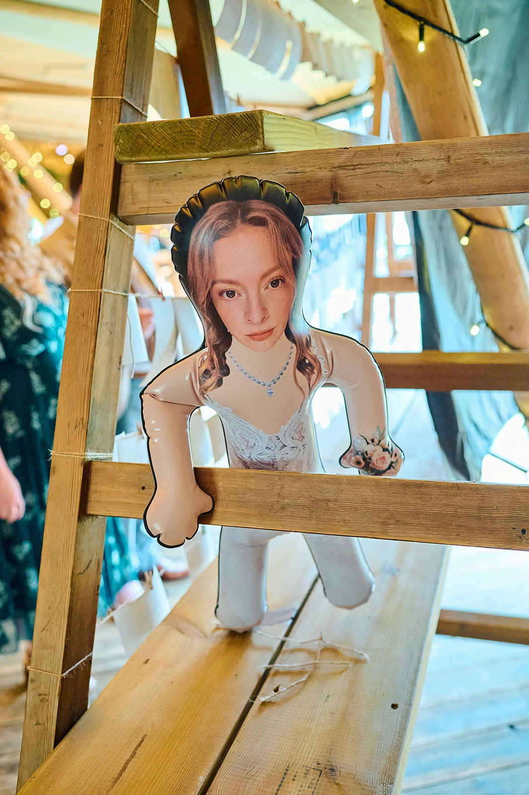 A quirky inflatable cut-out doll featuring the bride's face is propped up against wooden beams inside a brightly lit tipi wedding venue.