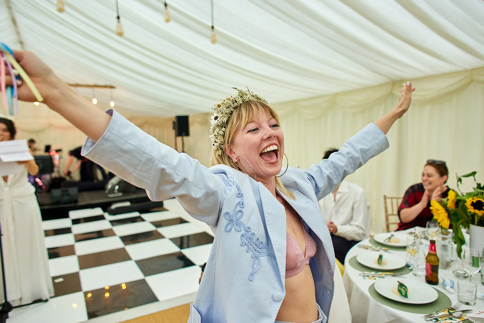 A joyful bride wearing a light blue embroidered suit jacket and a flower crown raises her arms with pure laughter inside a marquee wedding reception.