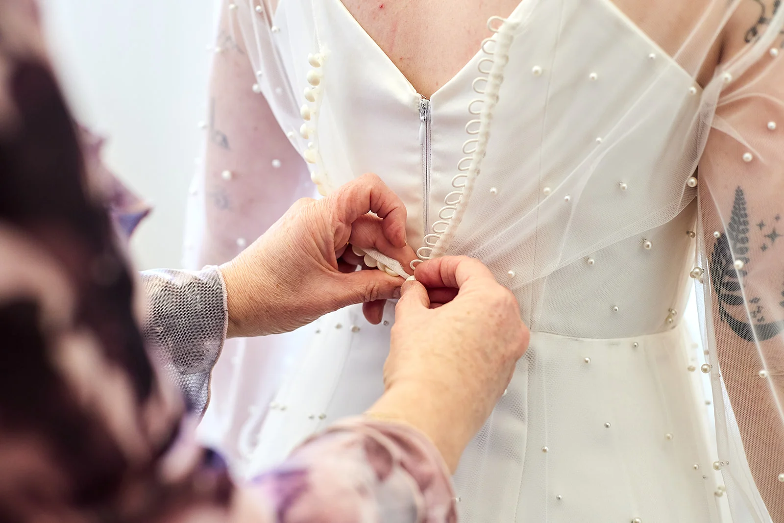 A close-up shot captures hands carefully fastening the delicate button-loop closure on the back of a pearl-embellished wedding dress during bridal preparations.