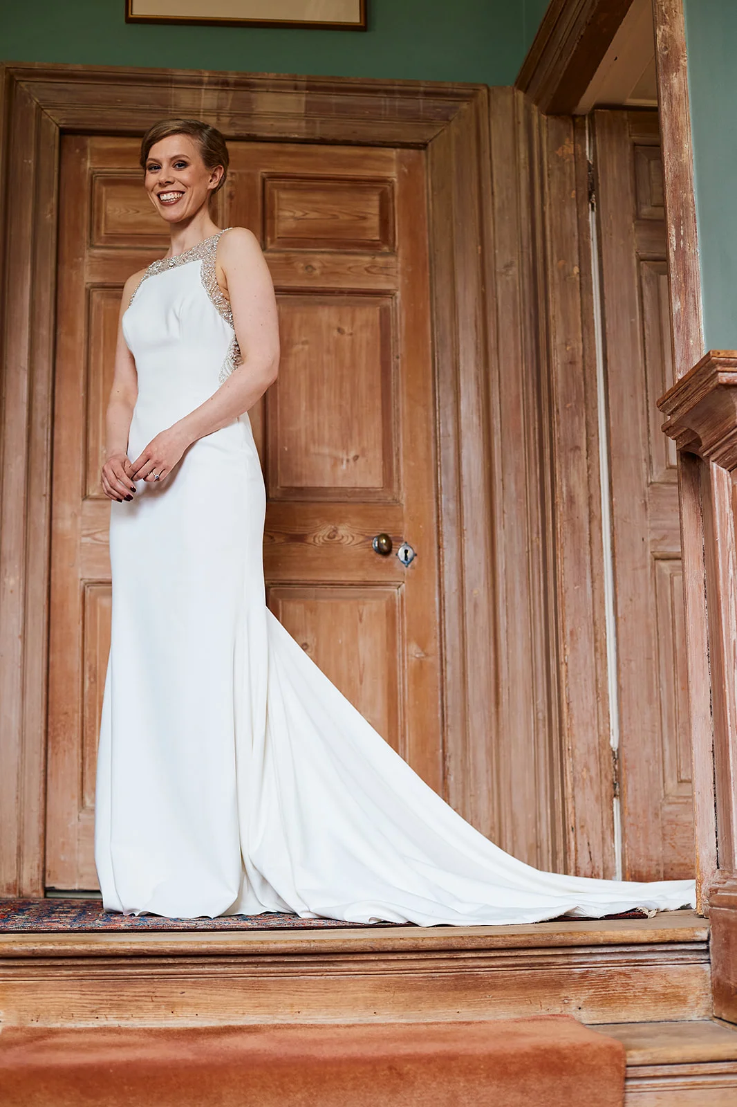 A smiling bride wearing a crepe column wedding dress with an embellished high neckline and a sweep train poses on a wooden staircase indoors.