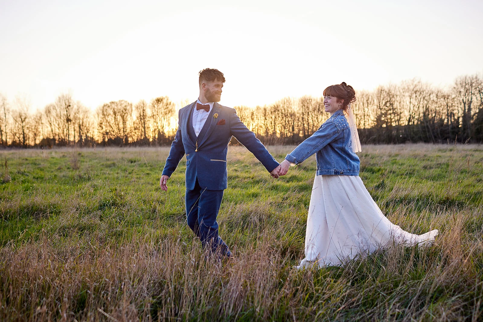 A newlywed couple walk hand in hand through a grassy field during golden hour, smiling at each other as the sun sets behind the tree line.