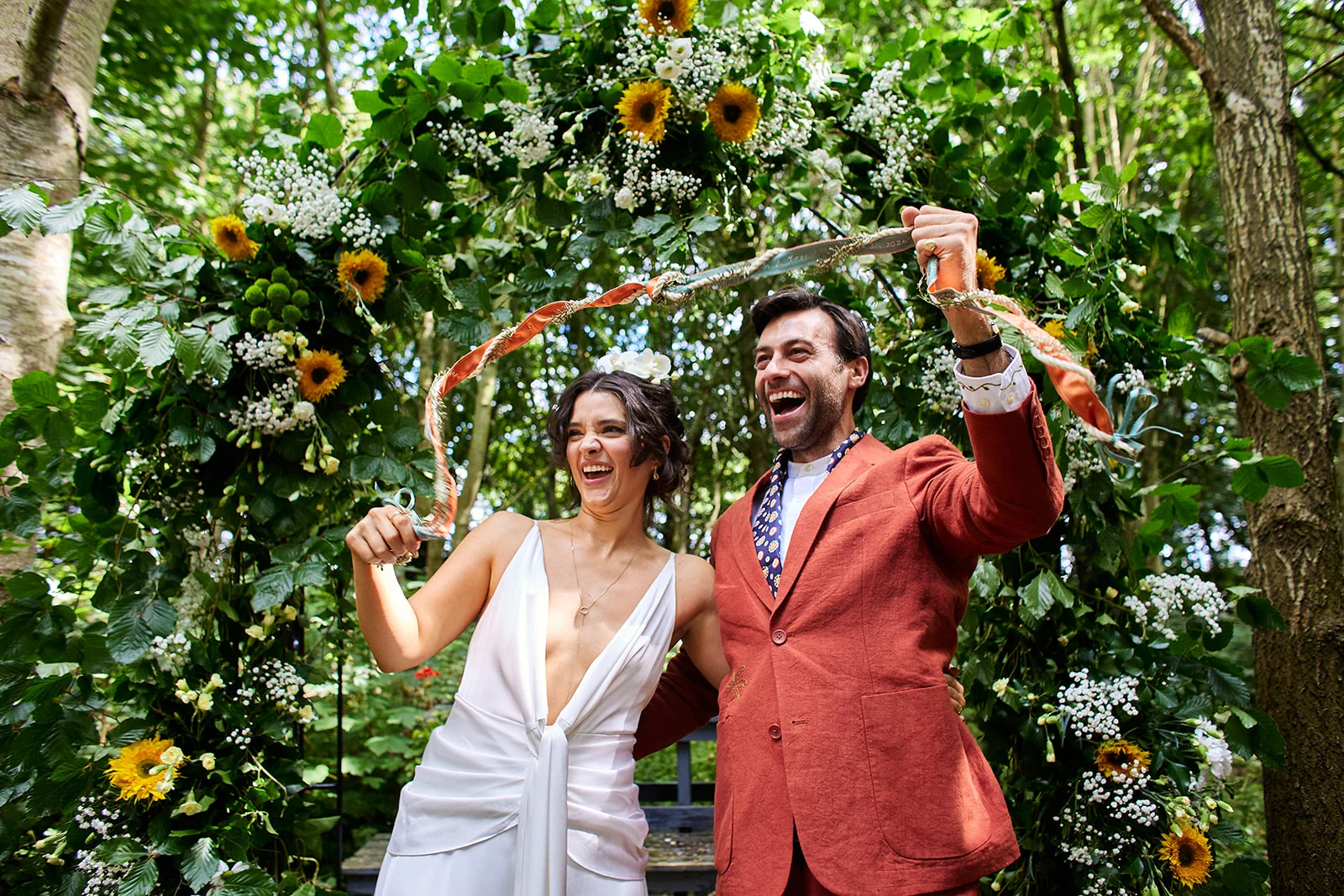 A joyful newlywed couple laughs beneath a lush floral ceremony arch adorned with sunflowers and foliage in a woodland setting, holding up ribbons after a handfasting ritual.