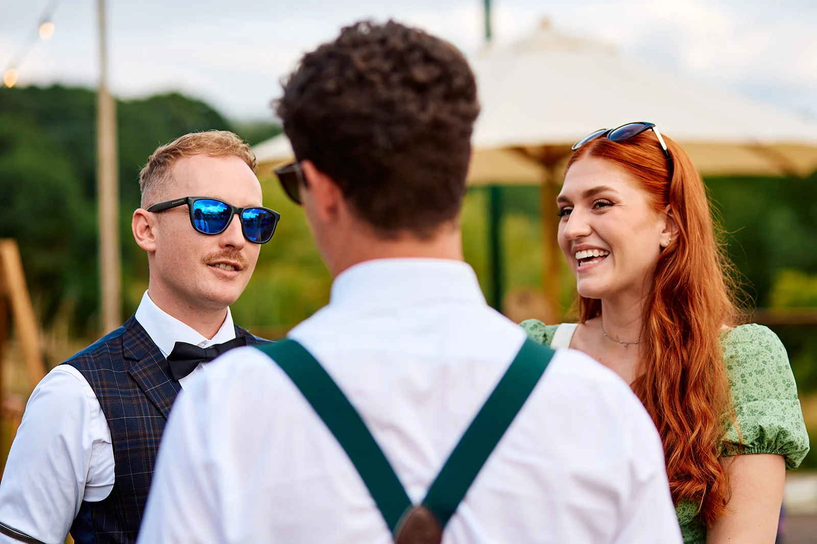 A smiling guest with red hair in a green dress and a guest wearing sunglasses and a waistcoat laugh and chat during an outdoor wedding drinks reception.