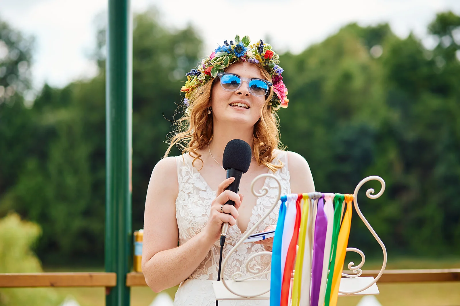 A bride wearing mirrored sunglasses and a vibrant floral crown delivers her speech outdoors while holding a microphone at a festival-themed wedding.