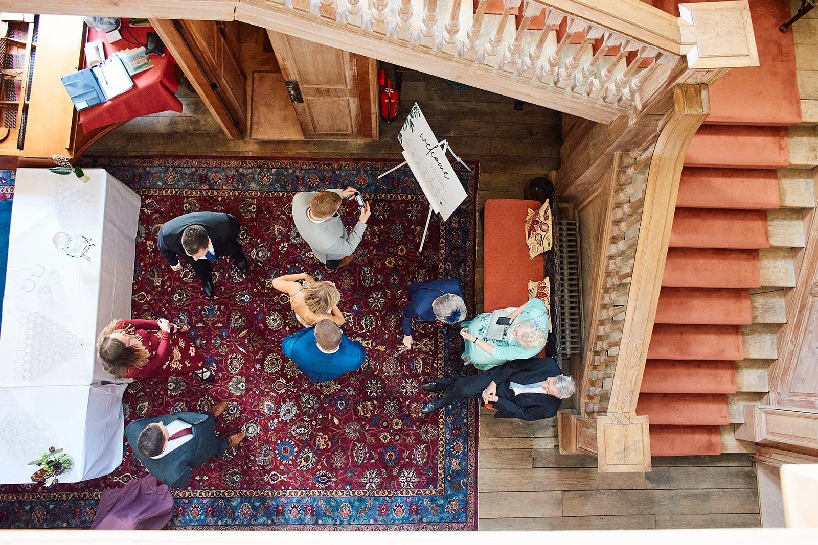 An elevated, high-angle view captures wedding guests mingling near a grand wooden staircase and an ornate patterned rug inside a historic venue.