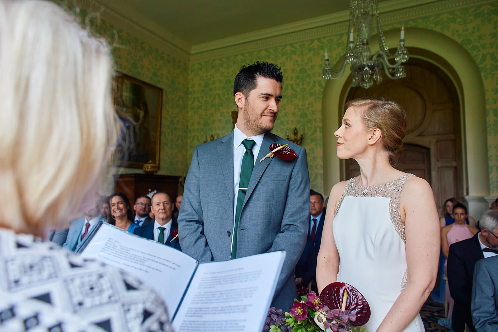 A bride and groom exchange vows during an indoor civil ceremony in an elegant country house, with a celebrant officiating in the foreground.