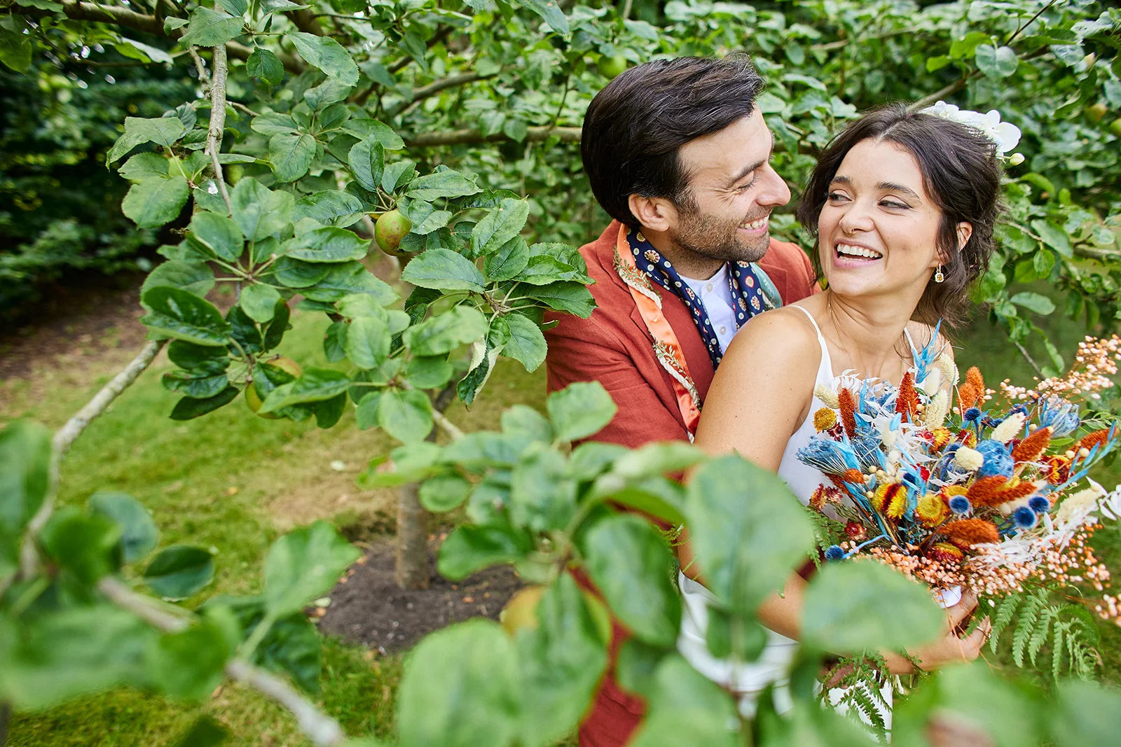A joyful couple embraces happily amidst an apple orchard, with the bride holding a colourful dried flower bouquet while looking back lovingly at the groom.
