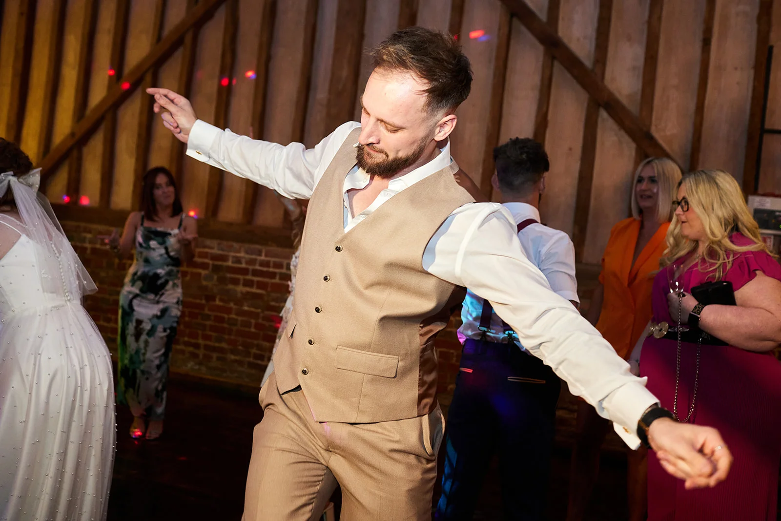 A groom wearing a tan waistcoat and open-collared white shirt dances enthusiastically on a packed dance floor inside a rustic barn wedding venue.