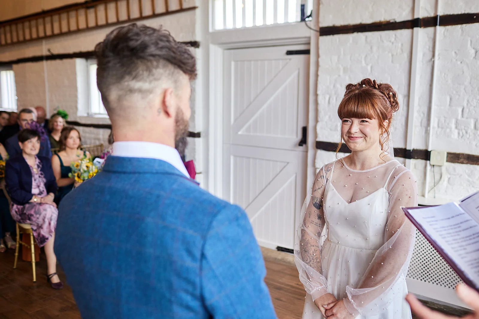 A groom in a blue checked suit stands facing his smiling bride during their indoor civil ceremony inside a white brick barn.