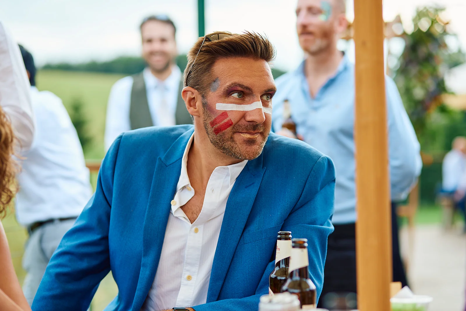 A wedding guest wearing a blue suit and festive face paint looks away smiling during an outdoor celebration.