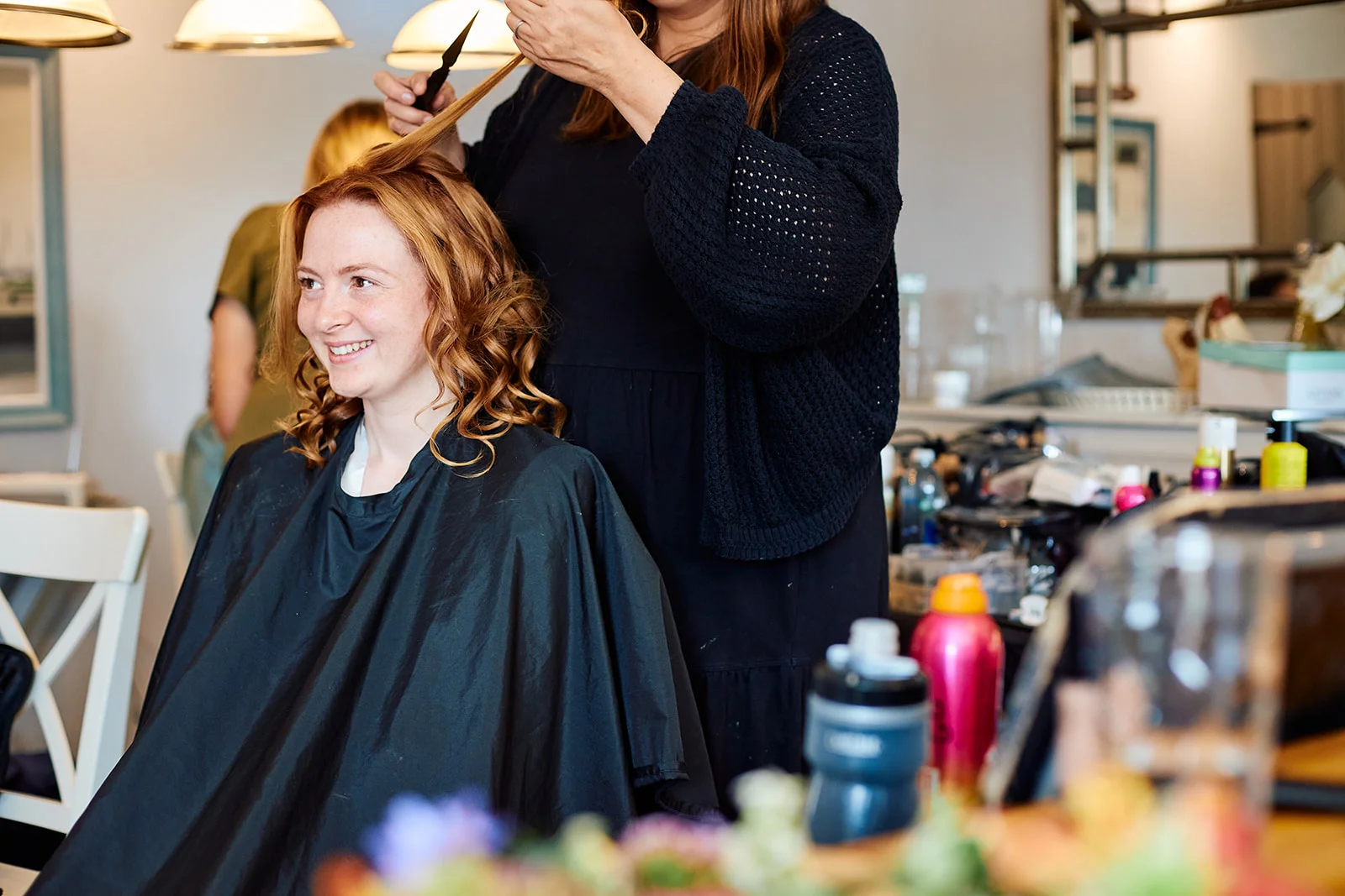 A smiling bride with red hair sits in a salon chair as a stylist styles her hair during morning bridal preparations.