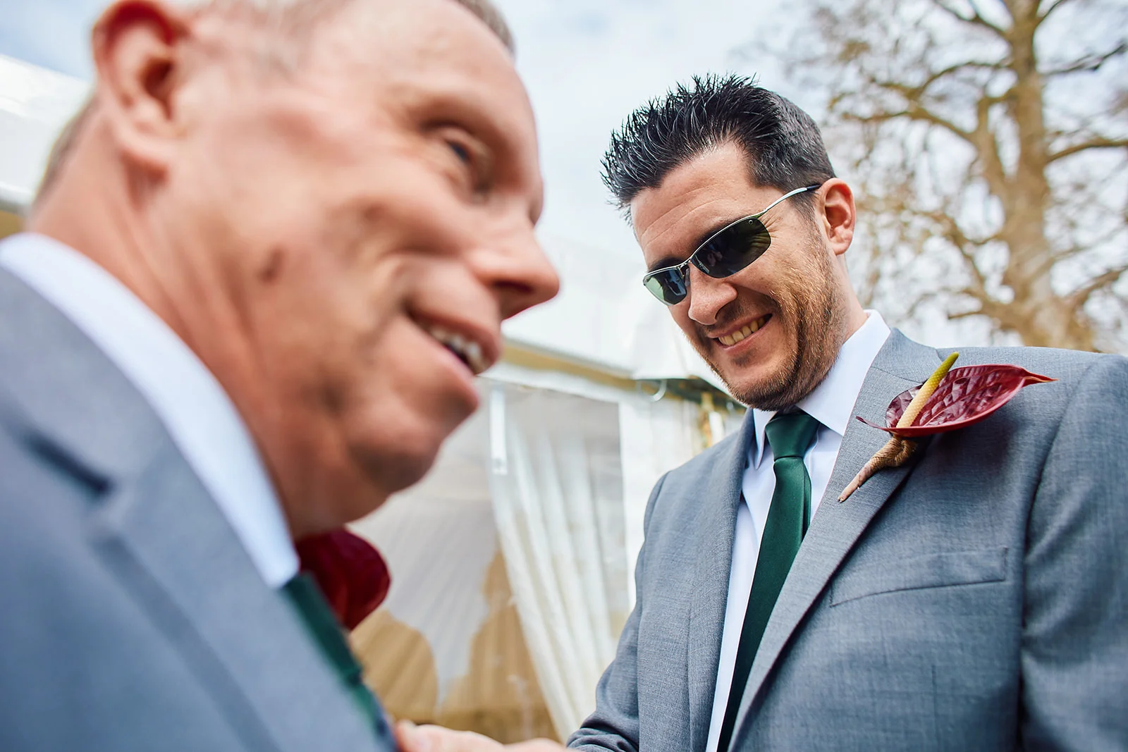 A guest in a grey suit and green tie wearing sunglasses smiles outdoors at a wedding celebration.