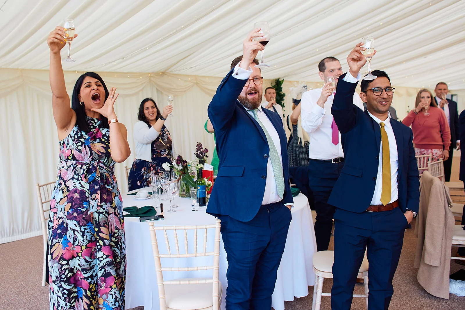 Excited wedding guests raise their glasses in a toast during speeches inside a brightly lit marquee.