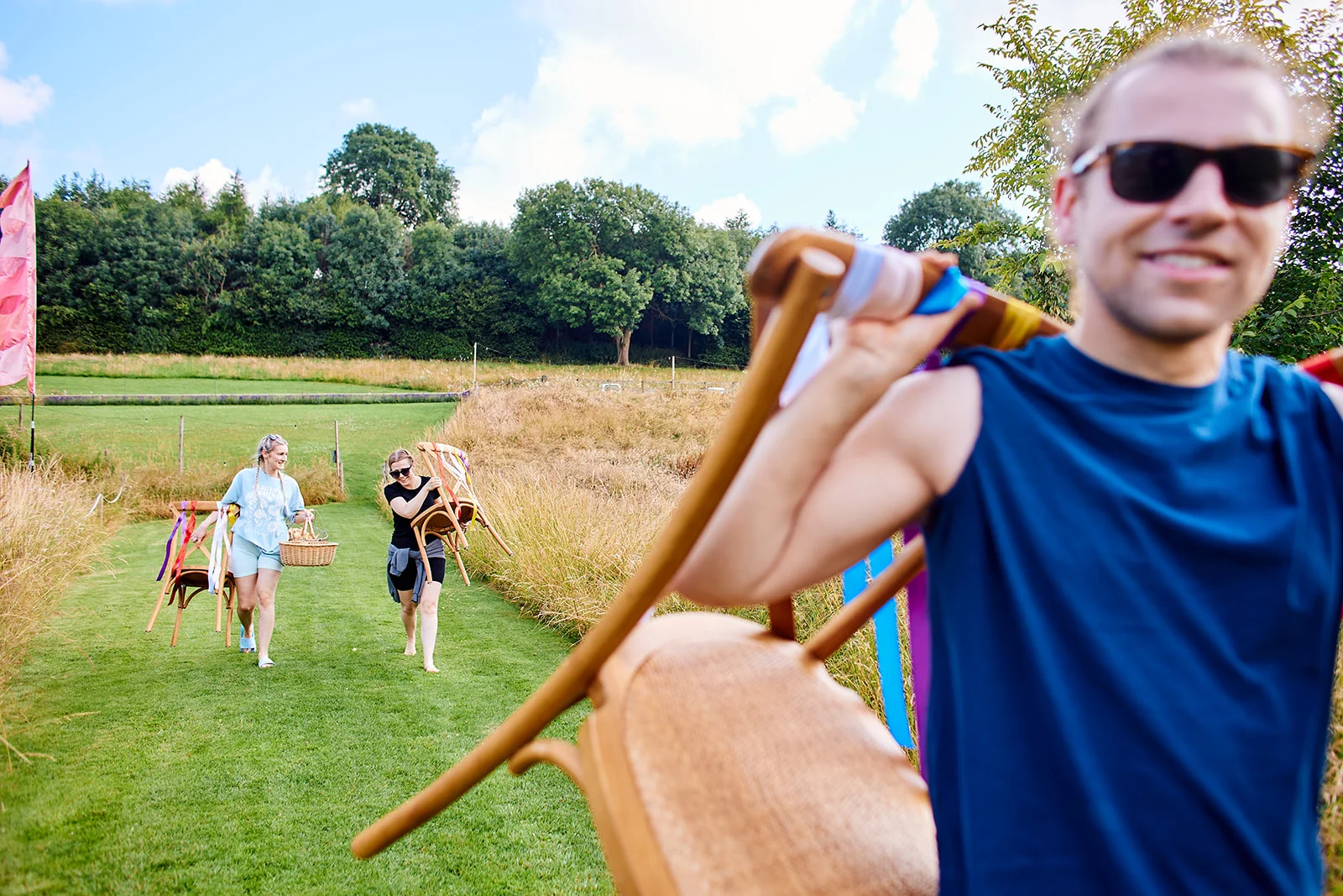 Guests joyfully carry chairs across a sunny green meadow during preparations for an outdoor festival style wedding.