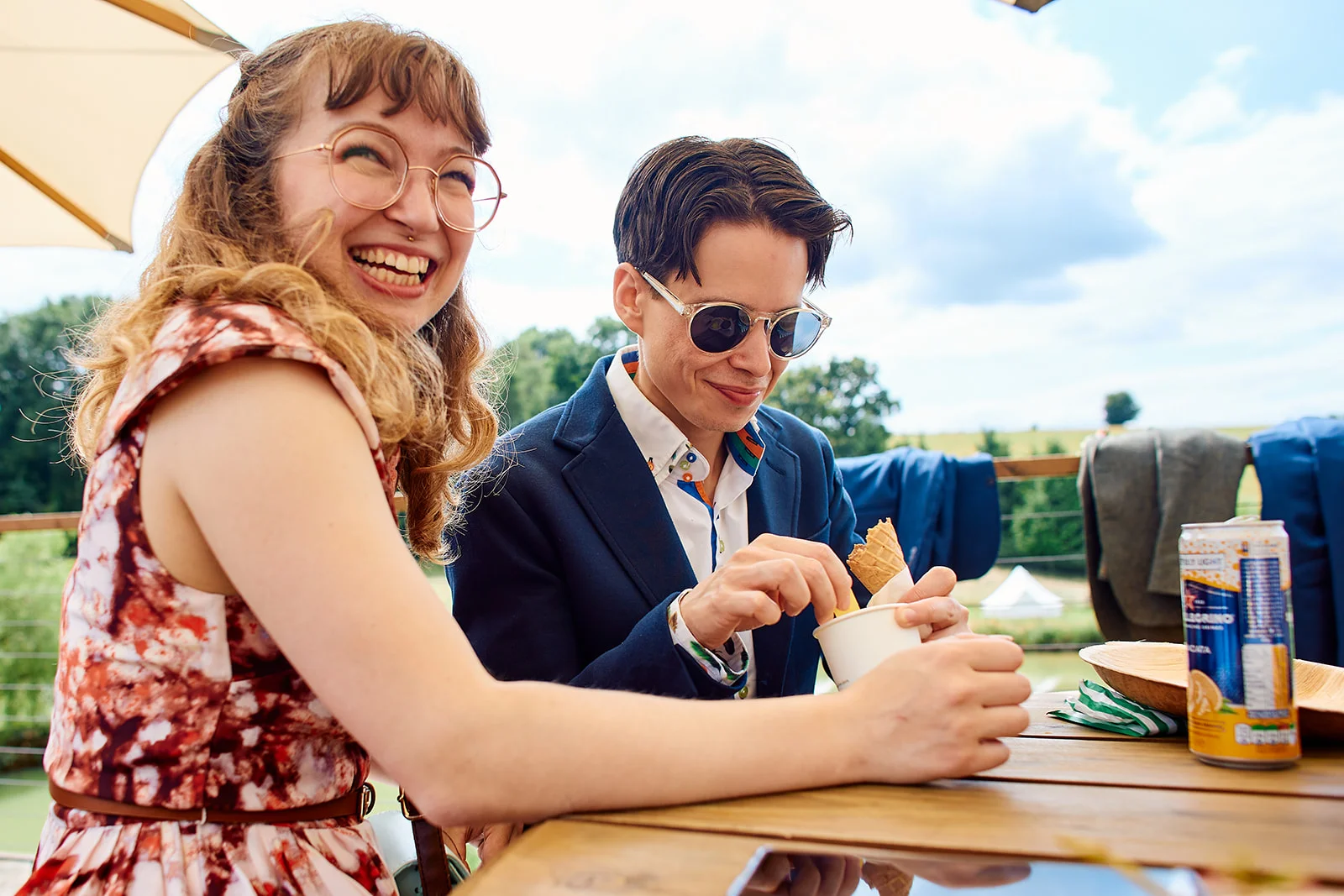A joyful same-sex couple shares a laugh outdoors on a sunny summer day while enjoying ice cream at a wooden table.