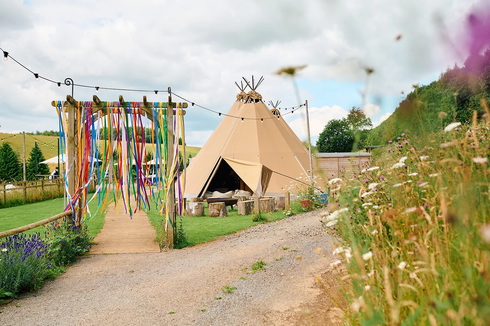 An outdoor festival wedding scene featuring a large tipis tent set against rolling green hills, with a colourful ribbon archway over a wooden walkway and wildflowers in the foreground.