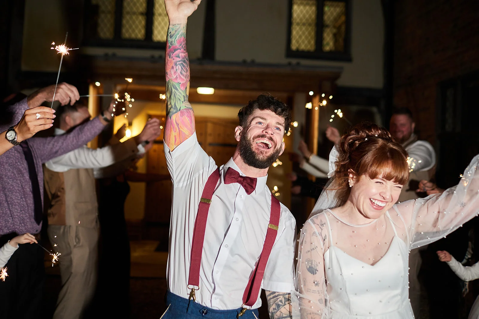A joyful newlywed couple laughs happily amidst a glowing sparkler tunnel with guests cheering at night.