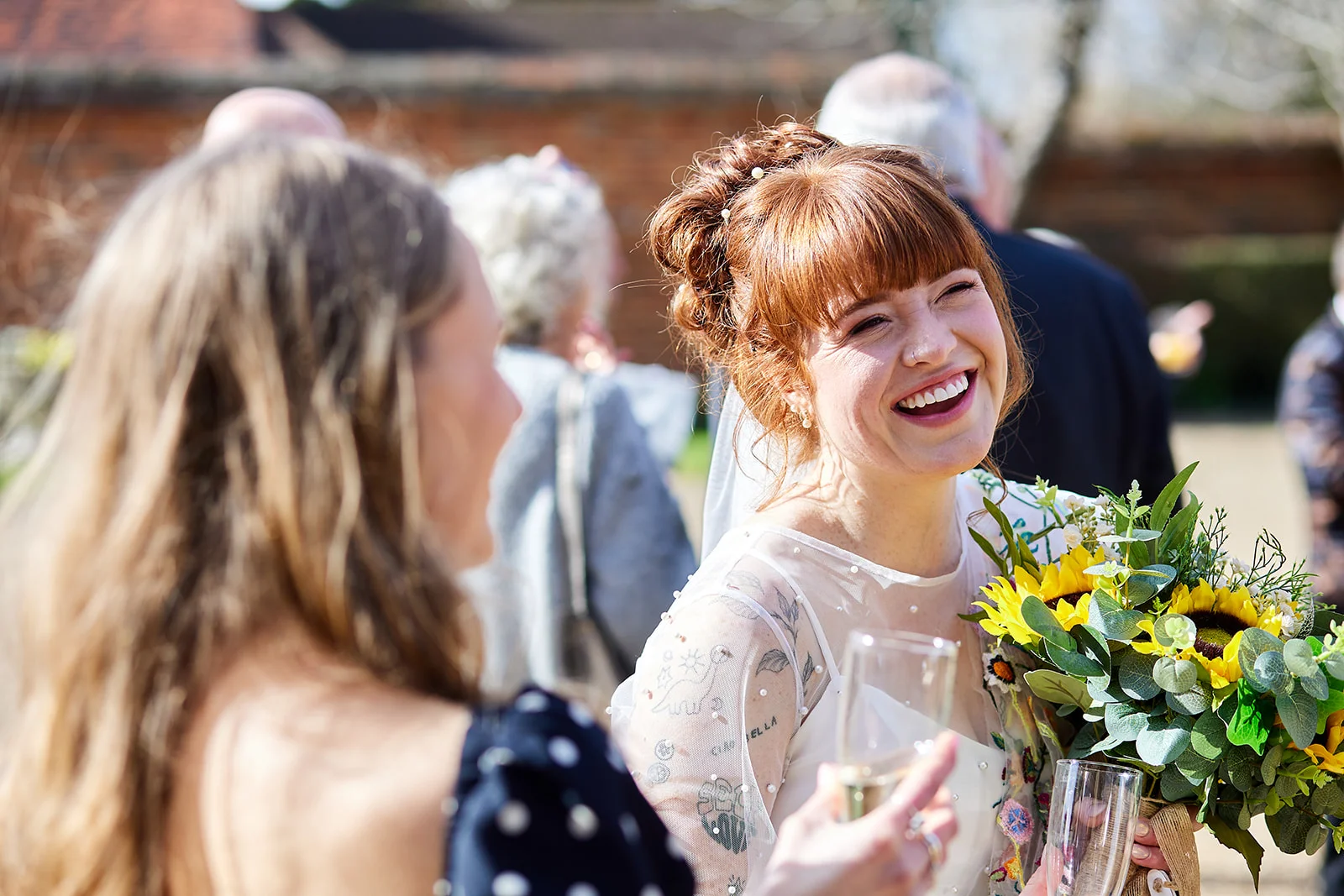 A smiling bride with red hair and tattoos laughs joyfully while holding a sunflower bouquet during an outdoor wedding drinks reception.