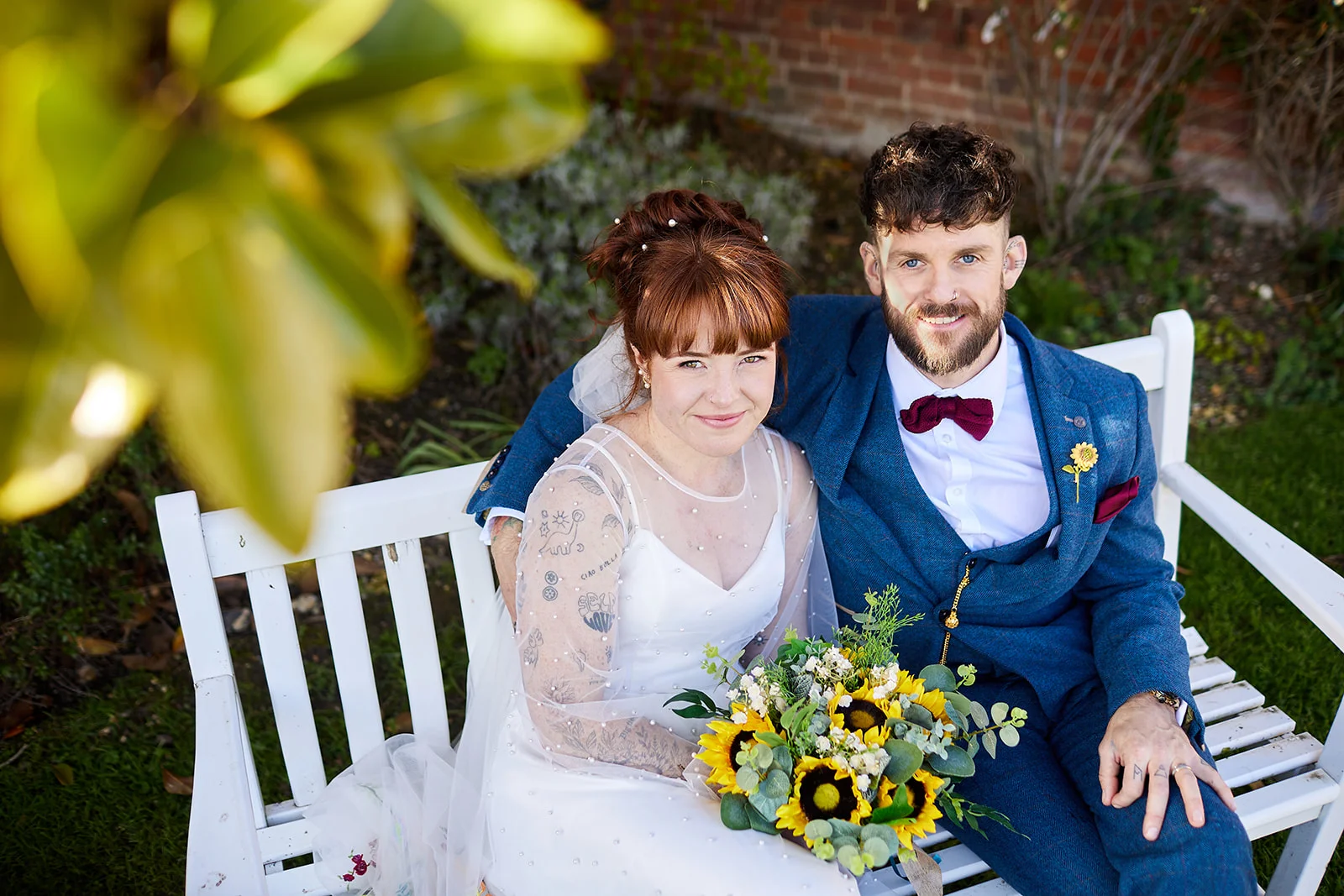 A newlywed couple sits together on a white wooden bench outdoors, with the bride holding a vibrant sunflower bouquet and showing off arm tattoos while the groom wears a blue suit with a burgundy bow tie.