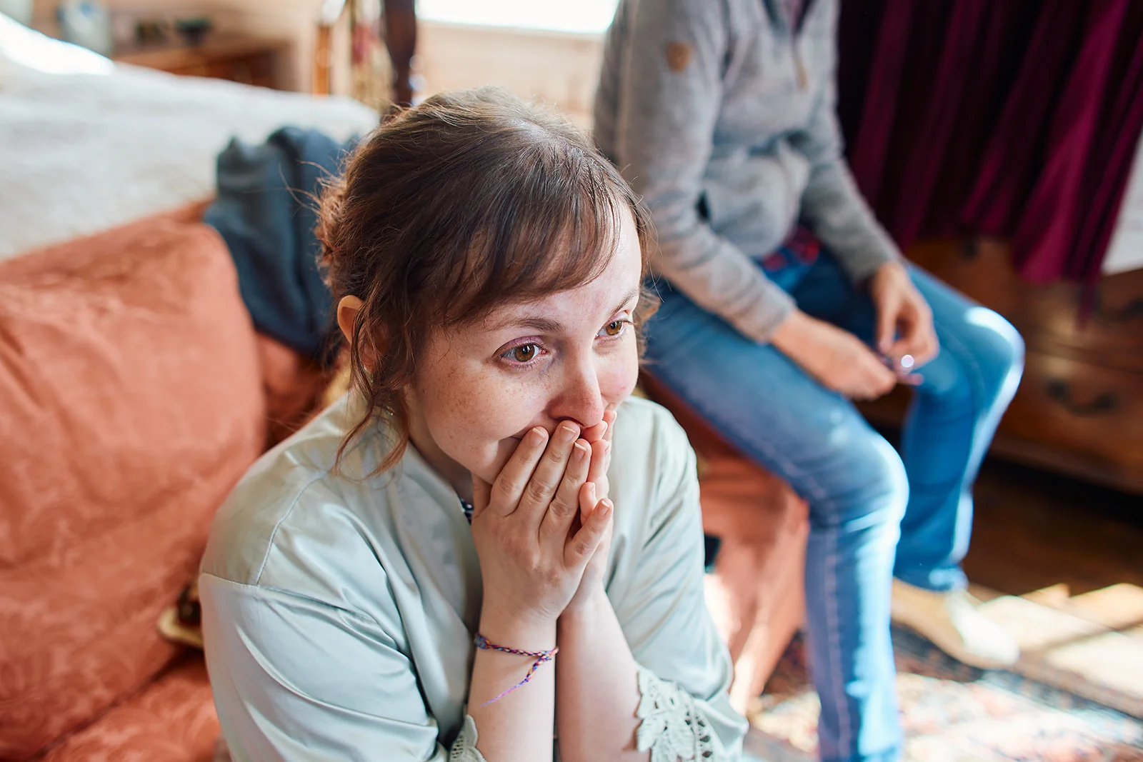 An emotional bride sits indoors wearing a robe with her hands covering her mouth in joyful disbelief while getting ready for her wedding.