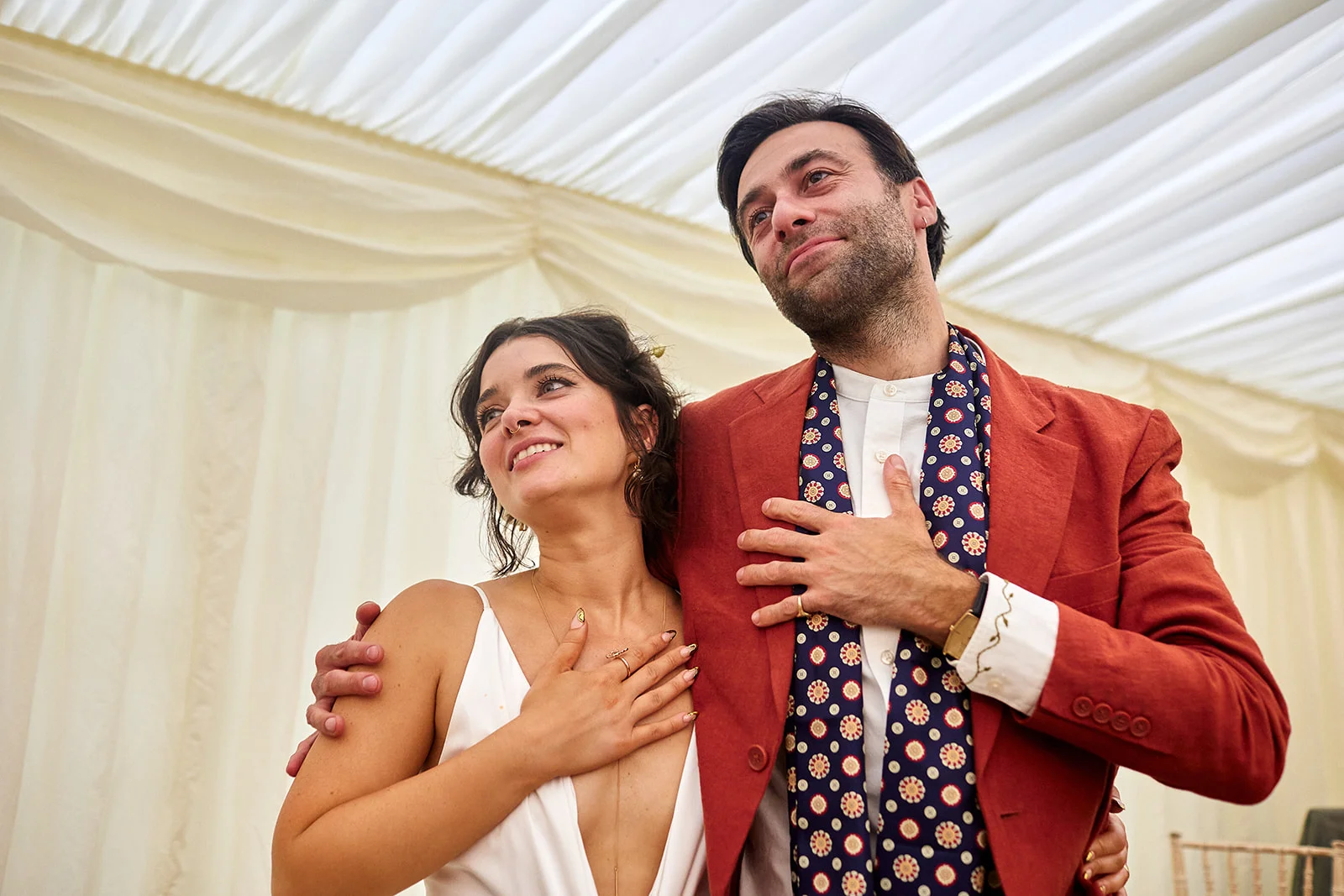 A joyful couple embraces under draped marquee ceiling lining, with the groom wearing a rust-coloured suit and patterned scarf and the bride in a white slip dress.