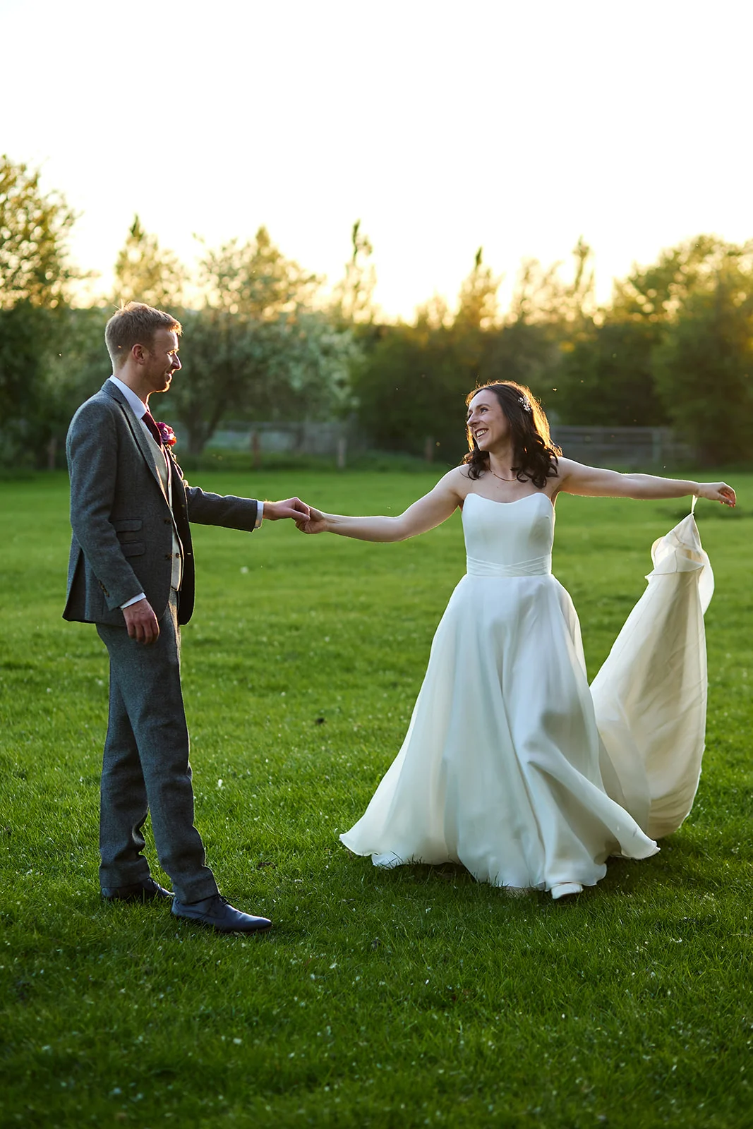 A bride and groom share a joyful moment holding hands and twirling in a lush green field during golden hour.