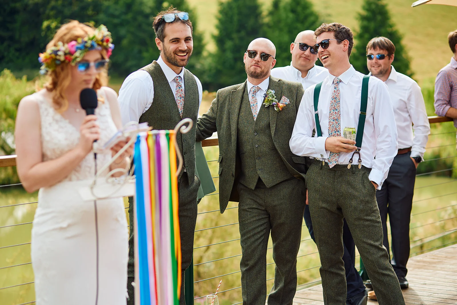 A bride wearing a floral crown delivers a speech outdoors while a group of smiling groomsmen in tweed waistcoats and sunglasses listen and laugh.