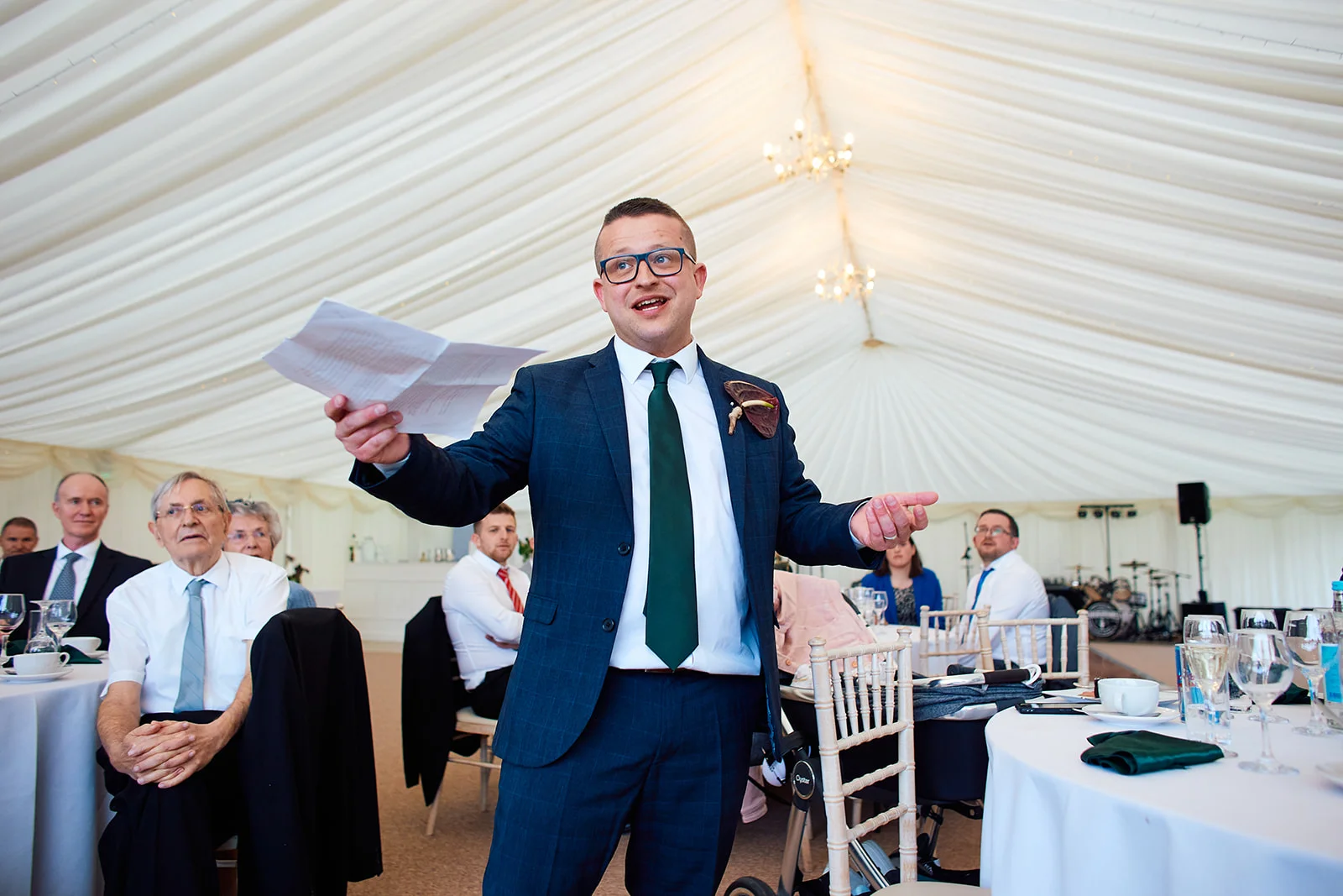 A smiling man wearing a navy suit and green tie delivers a speech inside a marquee, holding notes as guests listen from seated tables.