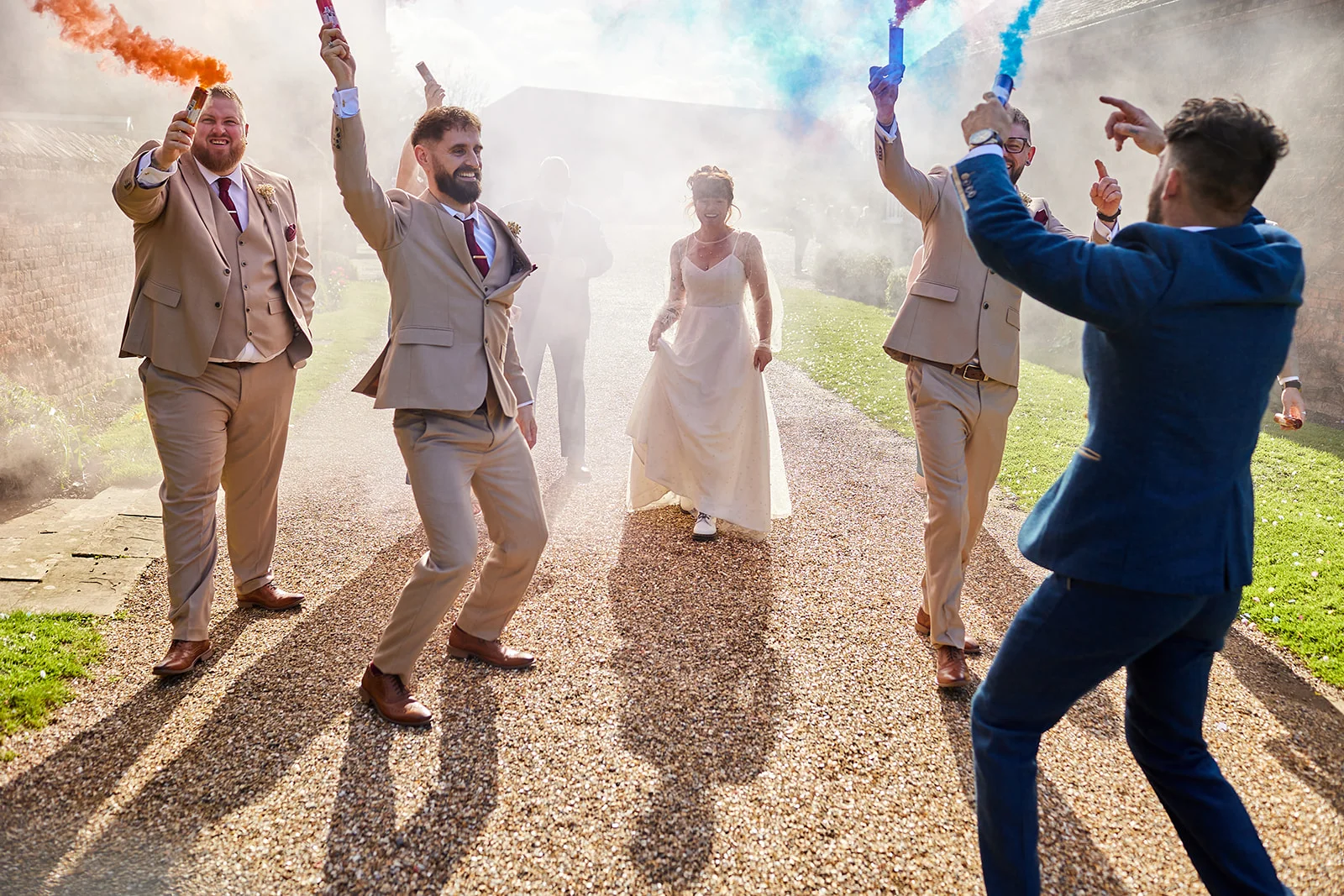 A joyous bride and groom walk down a gravel path outdoors, surrounded by groomsmen celebrating with colourful orange and blue smoke grenades.