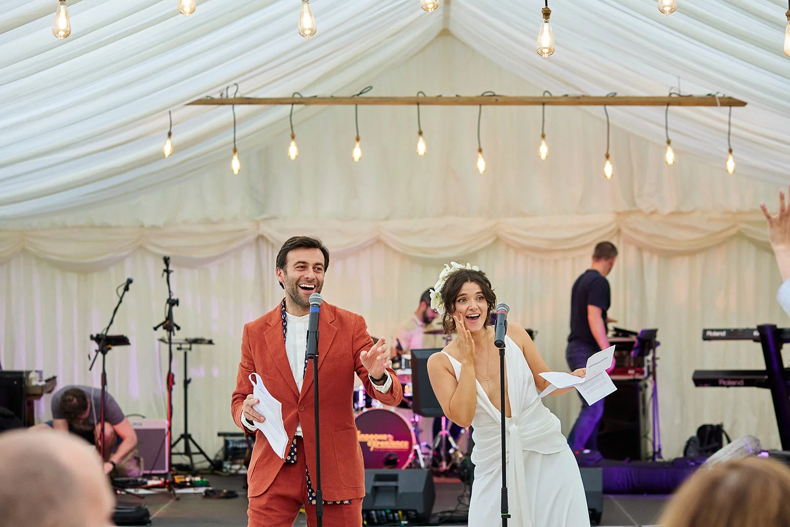 A joyful couple laugh and deliver speeches into microphones on a stage inside a draped marquee adorned with hanging festoon lights.