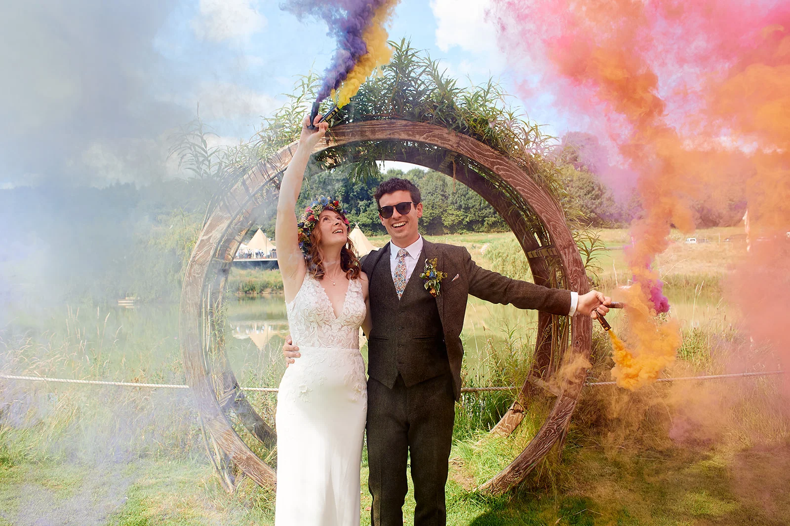 A joyful newlywed couple pose outdoors in front of a rustic wooden circular archway by a lake, joyfully setting off colourful smoke grenades in vibrant shades of blue, orange and pink.