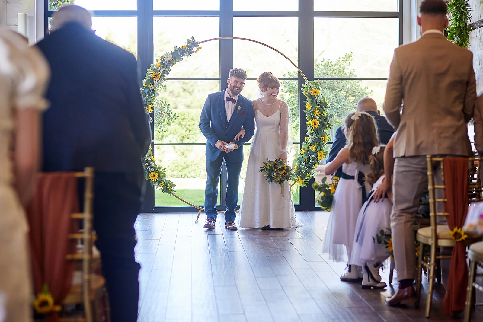 A smiling newlywed couple stands together under a floral circular arch adorned with sunflowers during their light-filled indoor wedding ceremony.